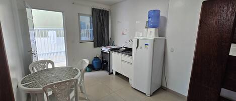 Een kleine keuken met een koelkast, spoelbak en waterdispenser. Er is een eettafel met stoelen, een raam met gordijnen en een uitzicht op een balkon.