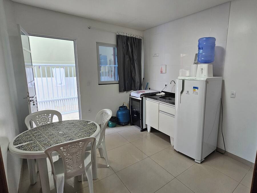 A compact kitchen with a refrigerator, sink, and a water dispenser. There's a dining table with chairs, a window with curtains, and a view of a balcony.