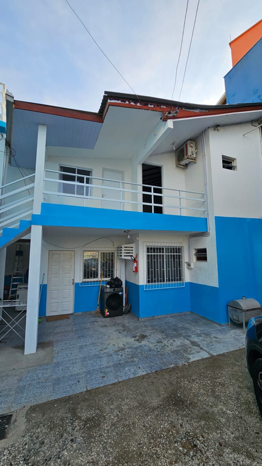 Um edifício de dois andares com esquema de cores azul e branco, uma área externa coberta e um extintor de incêndio visível.