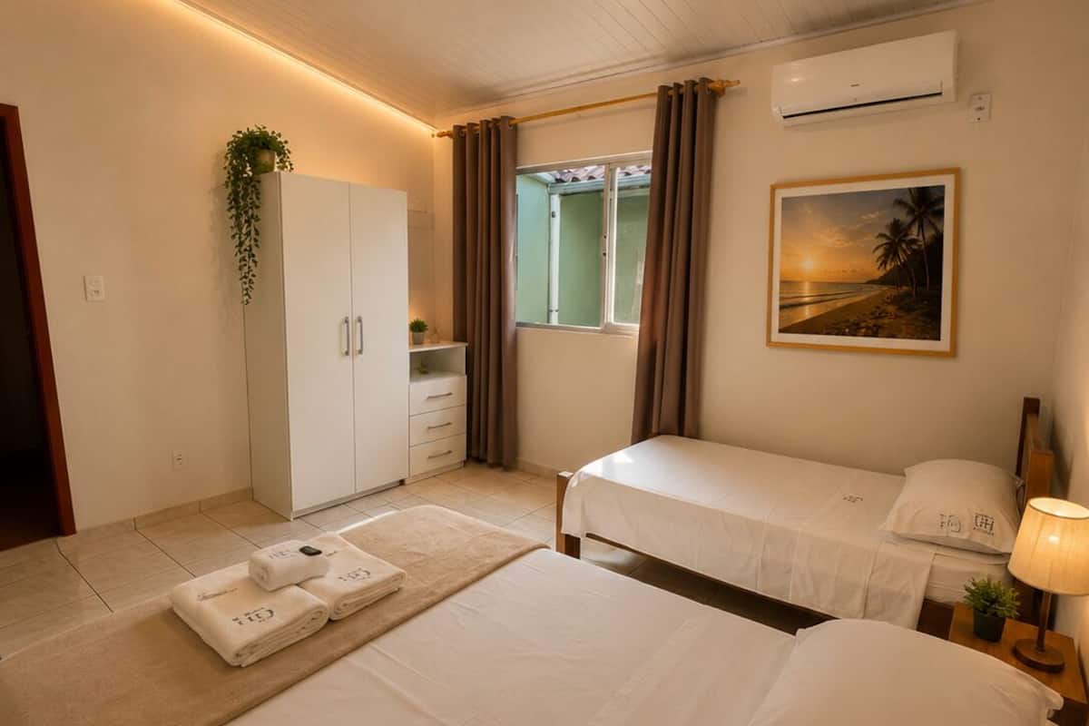 A hotel room with two beds, a wardrobe, a window with curtains, an air conditioner, a painting, and a plant.