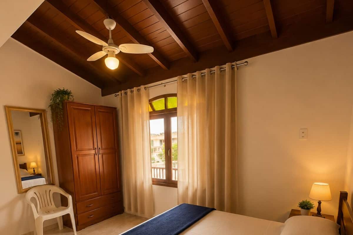 A hotel room with a bed, a ceiling fan, a window with curtains, a wooden wardrobe, a small table with a lamp, and a chair.