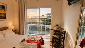 A hotel room with a bed, a desk, a TV, and a balcony with a view of buildings. - GH Barra da Lagoa (Florianópolis)