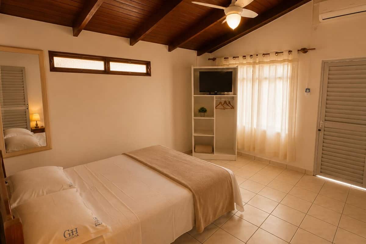 A bedroom with a bed, a nightstand with a lamp, a ceiling fan, a TV, and a window with curtains.