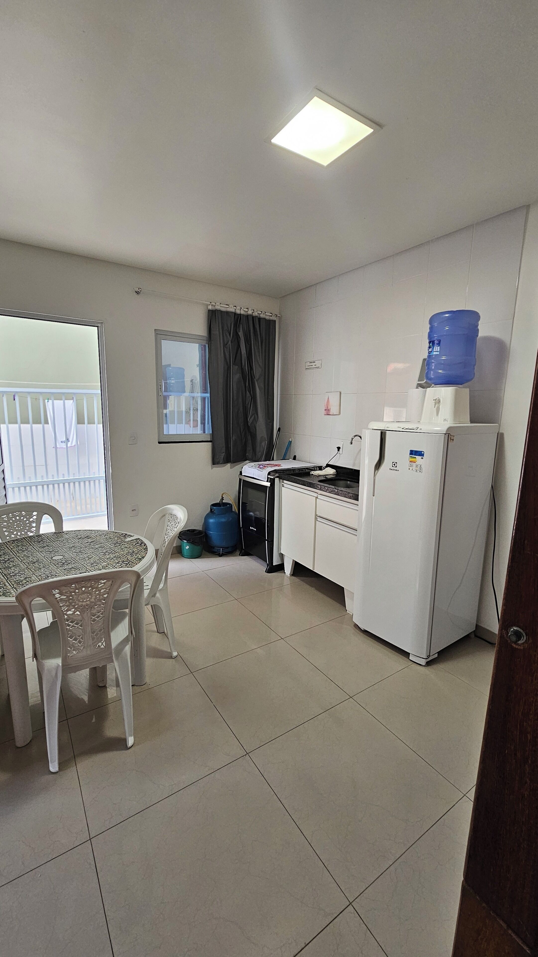 A small kitchen with a refrigerator, a water dispenser, a table with chairs, and a window with curtains.