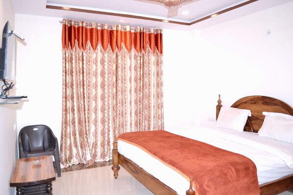 A bedroom with a wooden bed, a small table, a chair, and a curtain with a floral pattern. - Rudraksh Hotel & Restaurant (Kumbhalgarh)