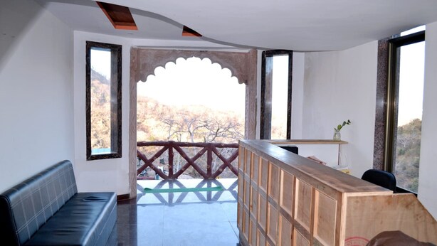 Une chambre d’hôtel moderne avec un comptoir d’accueil en bois, un canapé en cuir noir et une vue sur un paysage montagneux par une fenêtre en arc de cercle.
