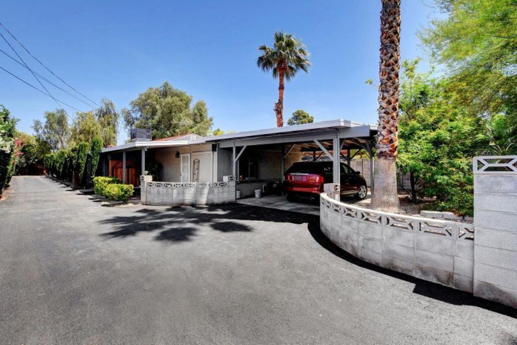 A modern house with a covered driveway, a red car parked inside, and a palm tree in the background.