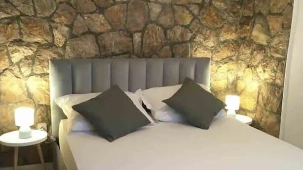Un dormitorio con pared de piedra, un cabecero tapizado en gris y dos mesitas de noche con lámparas.
