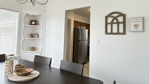 Un coin repas avec une table sombre, des chaises, un lustre et une cuisine visible à travers une porte ouverte.
