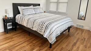 Un lit bien fait, recouvert d’une couvre-lit à motifs, une table de chevet avec une lampe et une fenêtre avec des stores.