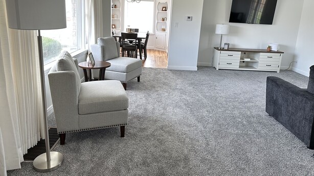 Un salon moderne avec un canapé gris, une table basse ronde et un fauteuil foncé. On y trouve également un coin repas agrémenté d’un lustre et d’un téléviseur à écran plat fixé au mur.