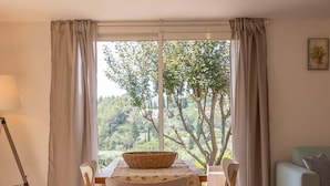 A dining area with a table and chairs, a basket on the table, and a view of trees and hills through large windows.