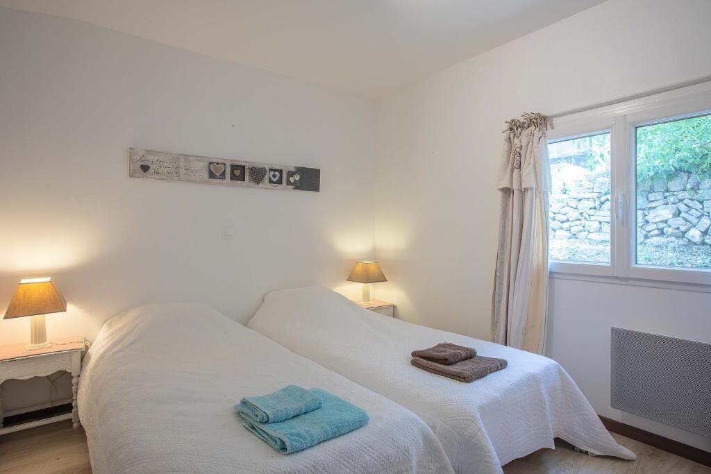Un dormitorio con dos camas, cada una con una colcha blanca y una lámpara en una mesita de noche. Hay una pieza decorativa sobre las camas y una ventana con cortinas en el lado derecho.