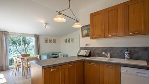 A modern kitchen with wooden cabinets, a countertop, and a dishwasher. There is a dining area with a table and chairs, and a window with curtains.