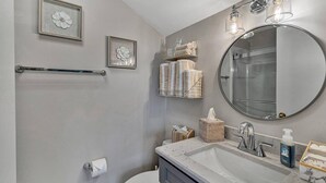 Ein Badezimmer mit Toilette, Waschbecken und Spiegel. An den Wänden befinden sich gerahmte Blumendekorationen, ein Handtuchhalter und ein Ablagebereich mit Handtüchern und Pflegeprodukten.