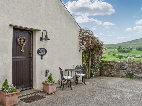 A cottage with a dark wooden door, a heart-shaped wreath, a sign for Stoney End Cottage, outdoor seating with two chairs and a table, and a stone wall with a sign. - A cosy cottage with spectacular views and optional hot tub. (Worton, near Leyburn)