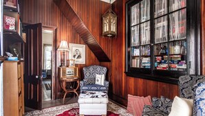 A cozy room with a patterned armchair, a matching ottoman, a red and white patterned rug, a wooden bookshelf, and a large window with newspapers. - Historical John Rast House (Roseburg)