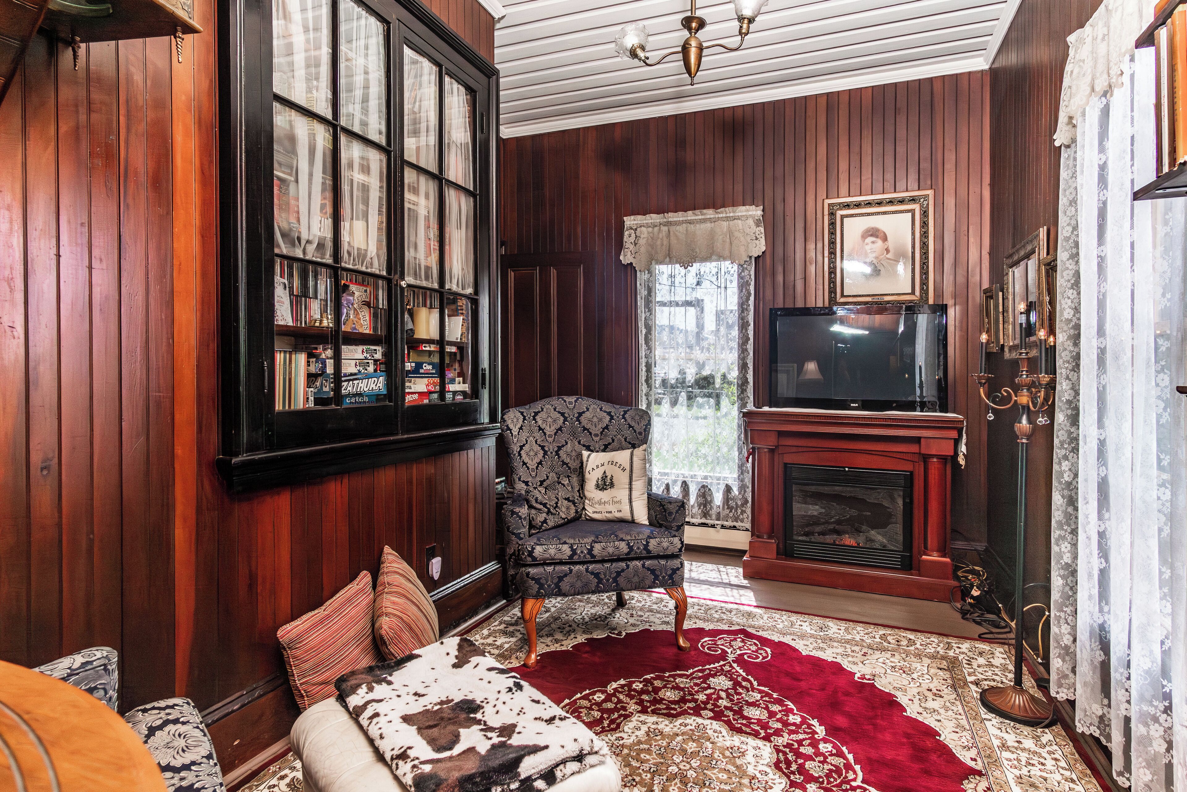 A cozy living room with a wooden paneled wall, a bookshelf, a patterned armchair, a red rug, and a fireplace.