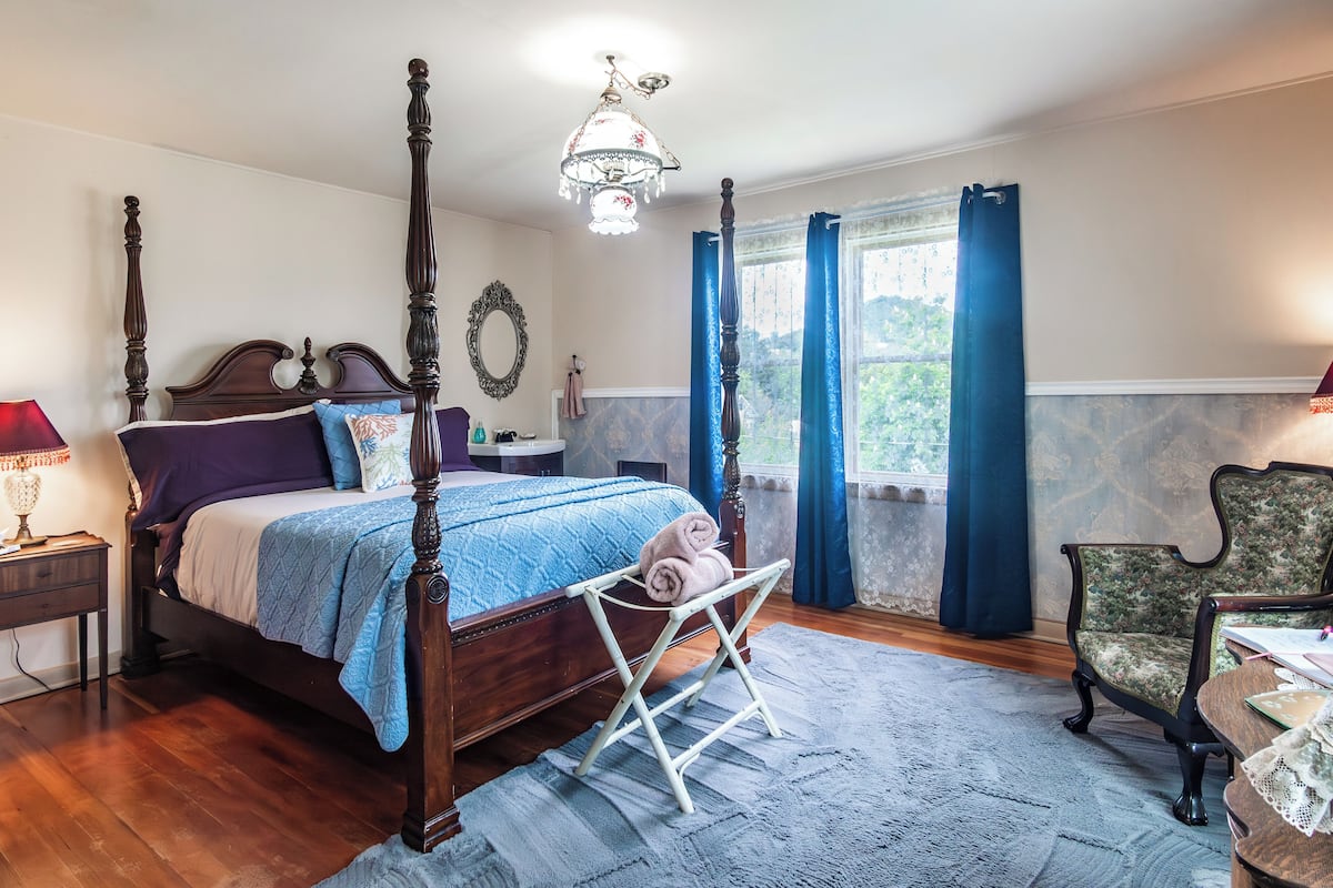 A four-poster bed with a blue bedspread, a wooden nightstand with a lamp, a chair with a floral pattern, and a view of the outdoors through a window with blue curtains.