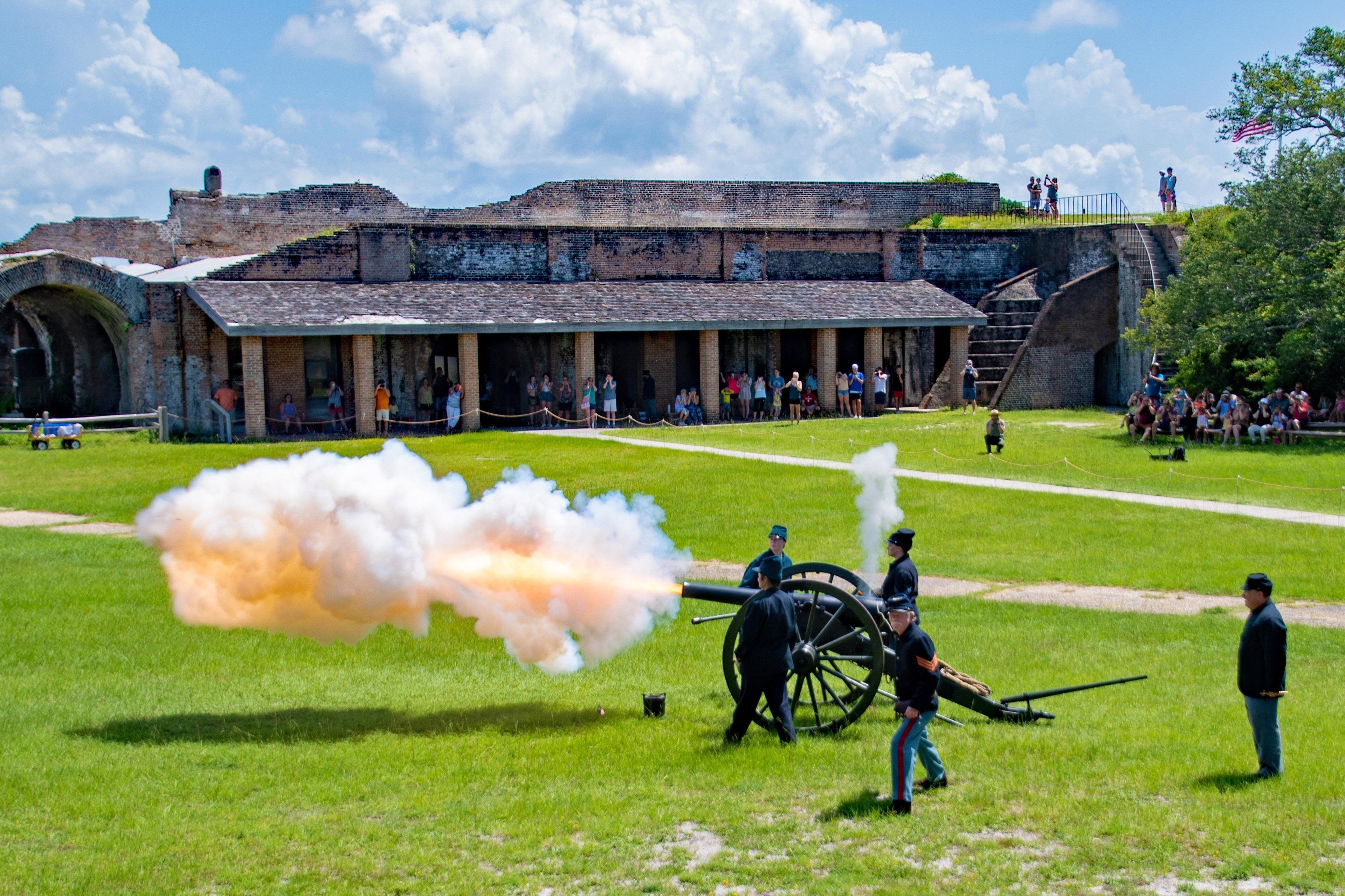A historical site with a large cannon firing, surrounded by a grassy field and a brick building with an arched entrance.