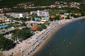 A coastal hotel with multiple buildings, a beach with numerous umbrellas, and a swimming area.