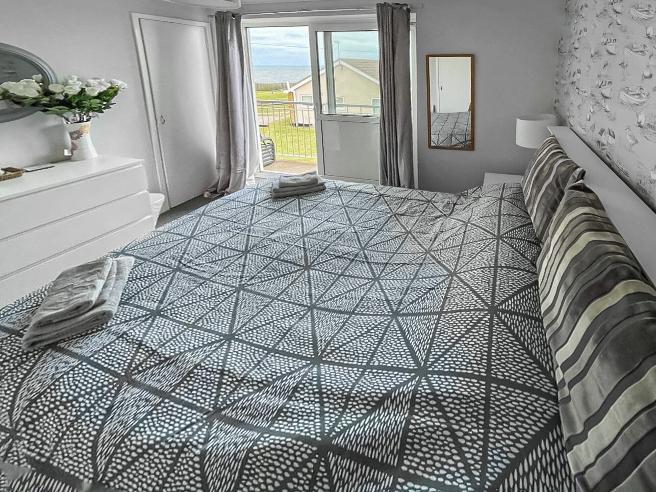 A bedroom with a geometric patterned bedspread, a bedside table with a vase of flowers, a mirror, and a view of the outdoors through an open door.