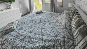 Un dormitorio con una colcha de diseño geométrico, una mesita de noche con un florero, un espejo y vistas al exterior a través de una puerta abierta.