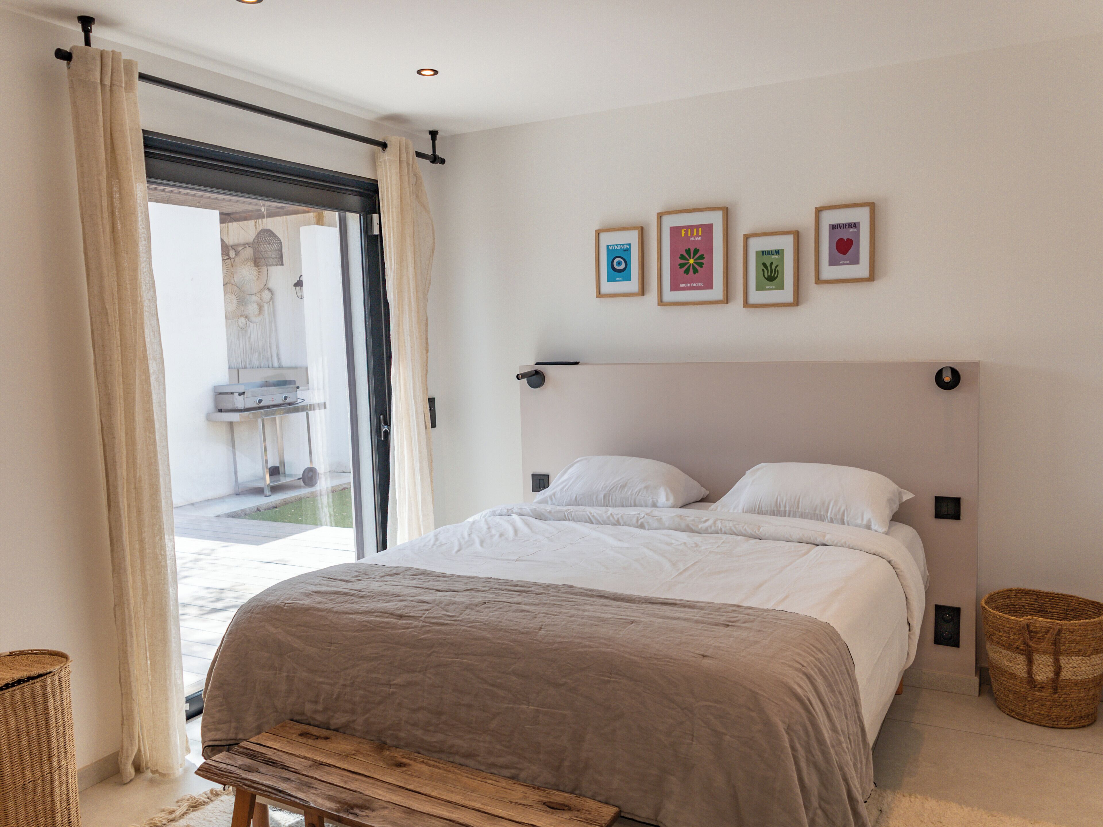 A bedroom with a bed, a bench, a wicker basket, and a view of an outdoor area.
