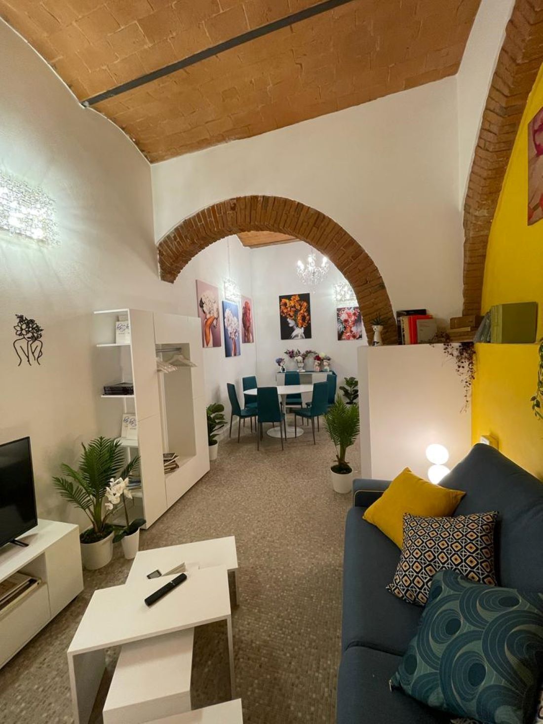 Un soggiorno con televisione, tavolino bianco e divano blu con cuscini a motivi decorativi. C'è una zona pranzo con tavolo e sedie, e un angolo cottura con pensile bianco e piante in vaso.