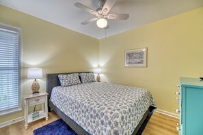 Un dormitorio con una cama, lámparas de noche, un ventilador de techo, un cuadro en la pared y una mesita de noche con una lámpara y una caja.