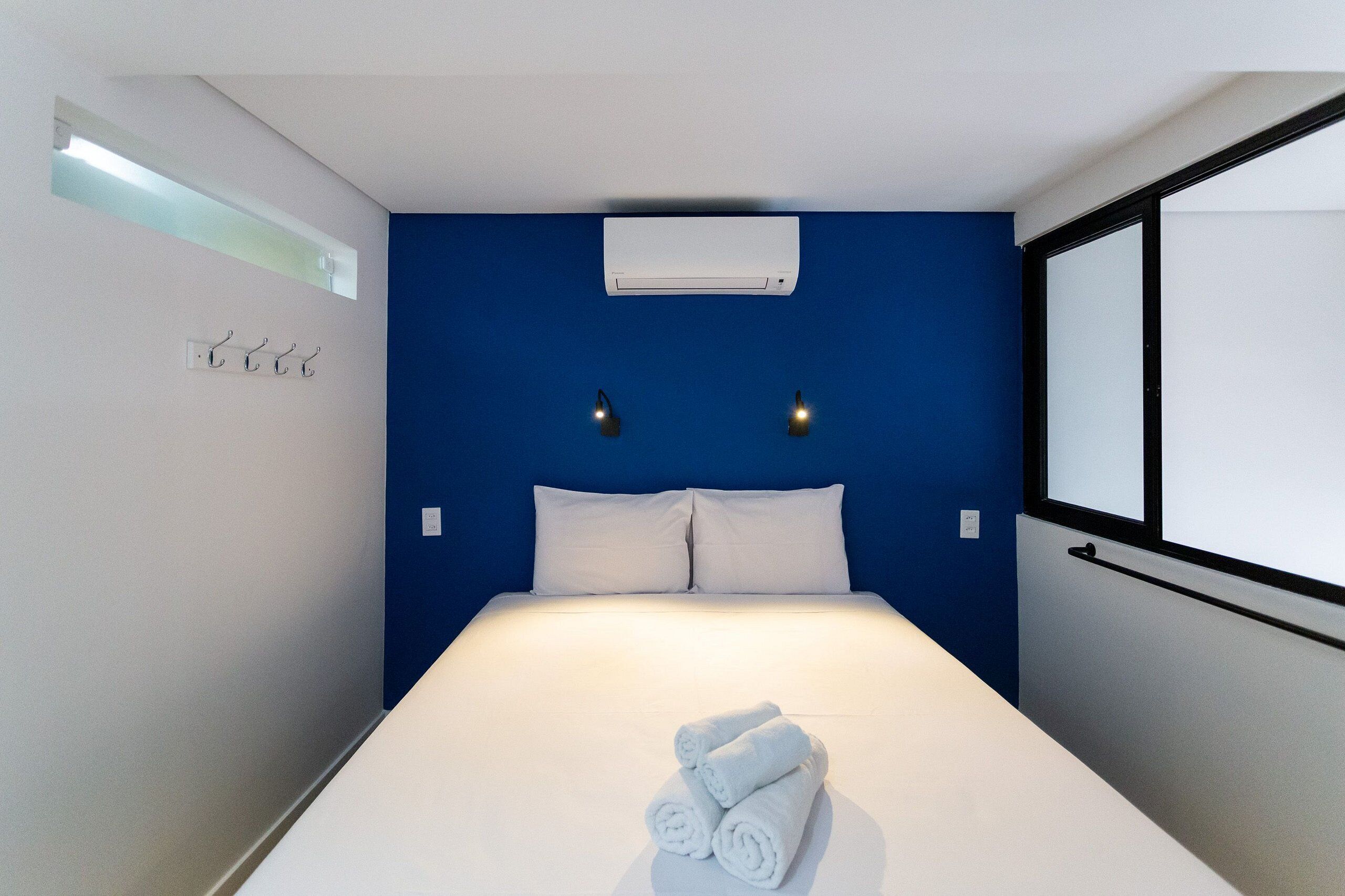 Une chambre d’hôtel avec un lit, du linge de lit blanc, un mur d’accent bleu, un appareil de climatisation et une fenêtre avec des stores.