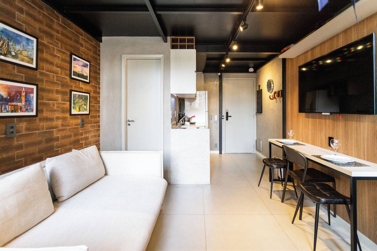 A modern hotel room with a sofa, a dining area with a table and chairs, a TV mounted on the wall, and framed pictures on the wall.
