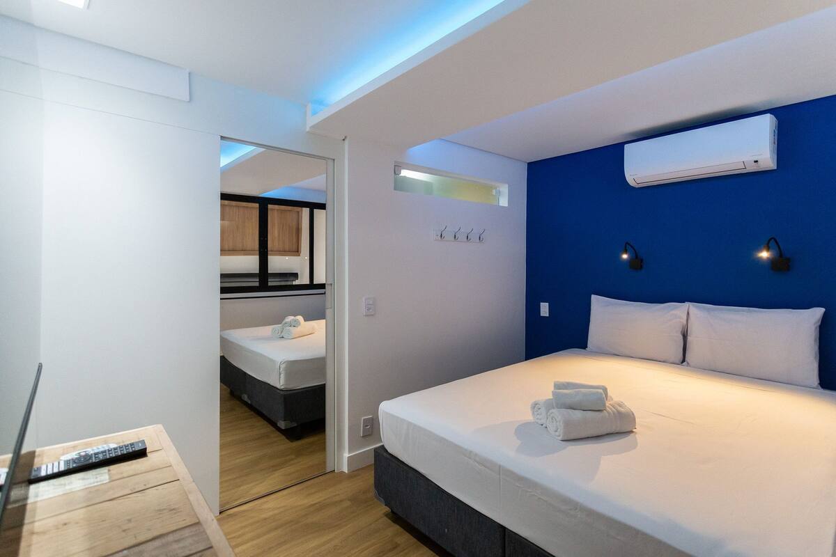 A modern hotel room with a large bed, a bedside table, a wall-mounted air conditioner, and a hallway leading to another room.