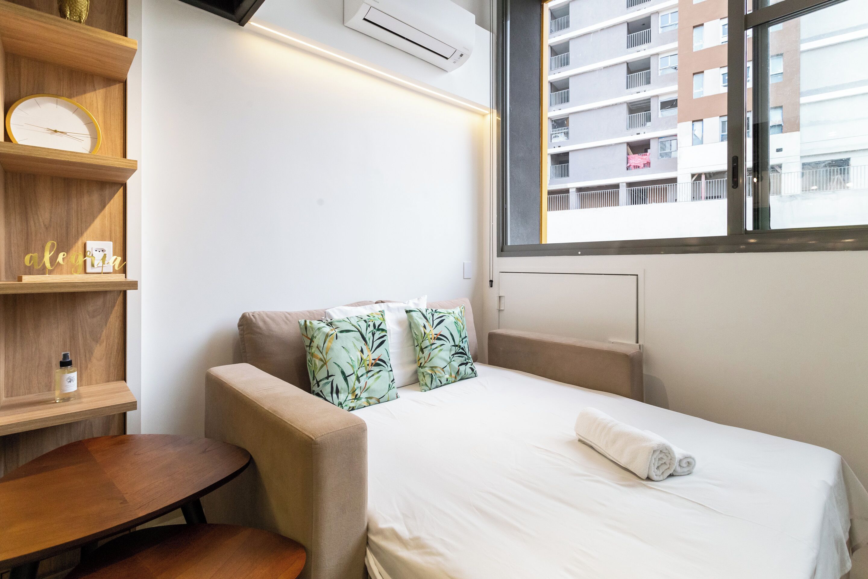 A hotel room with a bed, a small table, a sofa, a window with a view of buildings, and a shelf with a clock and a bottle.