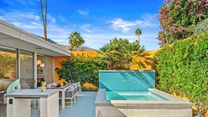 A modern outdoor pool area with a tiled deck, a lounge chair, and a dining table with outdoor seating. - The Desert Breeze at Sandpiper; Completely remodeled with private spa! (Palm Desert)