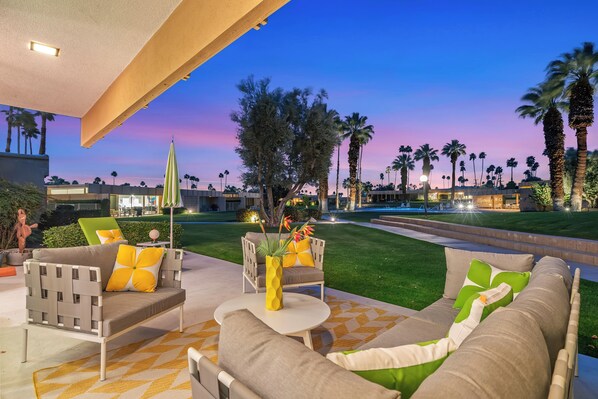 An outdoor seating area with a sofa, chairs, and a table, overlooking a well-manicured lawn and palm trees.