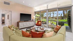 A modern living room with a large TV, comfortable seating, and a view of a well-maintained outdoor area. - The Desert Breeze at Sandpiper; Completely remodeled with private spa! (Palm Desert)