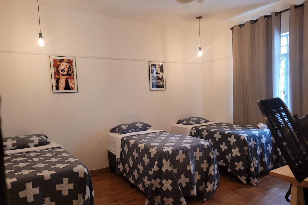 A room with three beds, each with a cross-patterned bedspread, a wooden floor, and two wall-mounted light fixtures.