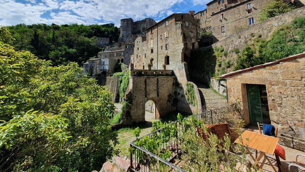A historic stone building with multiple levels and a prominent archway, surrounded by lush greenery and other traditional buildings. - Old ceramic factory close to Unesco itineraries (Sorano)