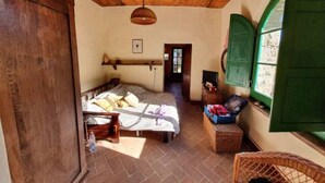 A rustic room with a wooden ceiling, a bed with a white bedspread, a wicker chair, and a small table with a basket. - Old ceramic factory close to Unesco itineraries (Sorano)
