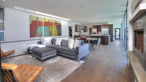 Un salon moderne avec un canapé d’angle gris, un fauteuil en cuir couleur sable et un coin repas avec une cuisine en arrière-plan.