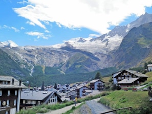 Uma vila de montanha com casas tradicionais, uma estrada visível e um hotel chamado "Hotel Alpenblick".