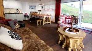 Ein gemütliches Wohnzimmer mit einem fellbezogenen Sofa, einem Holzesstisch und Blick auf einen grünen Hügel durch große Glastüren.