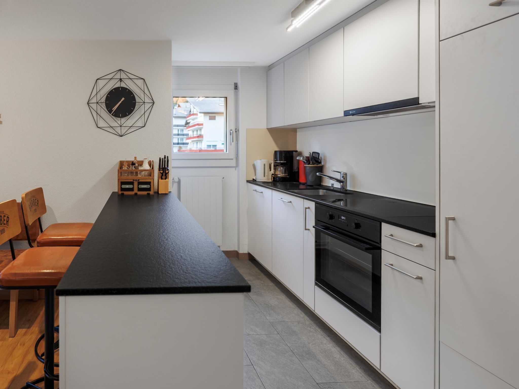 Cozinha moderna com bancada preta, armários brancos e forno embutido. Há uma janela com vista para edifícios.