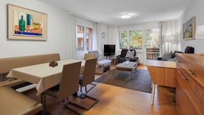 Een moderne woonkamer met een eethoek, een flatscreen televisie en een balkon met uitzicht op bomen.