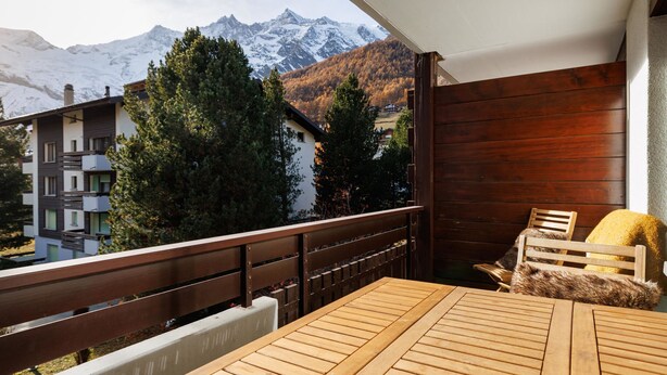 Ein Balkon mit Holztisch und Stühlen, mit Blick auf eine Berglandschaft mit schneebedeckten Gipfeln und herbstlich gefärbten Bäumen.