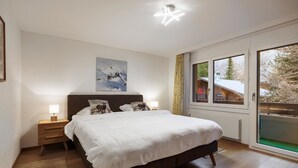Ein Schlafzimmer mit einem großen Bett, einem Nachttisch mit Lampe, einem Bild an der Wand und einem Fenster mit Blick auf Bäume.
