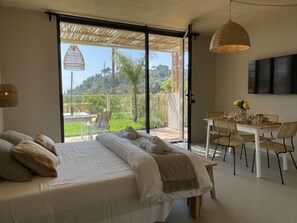 Un dormitorio con una cama grande, una zona de comedor con mesa y sillas, y un balcón con vistas a zonas verdes.