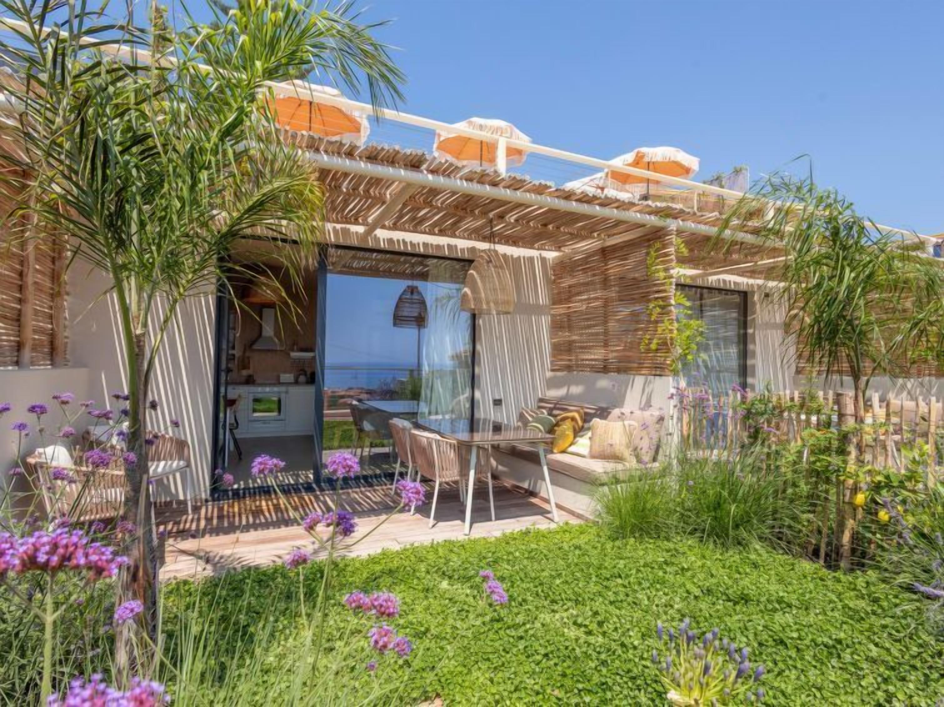 Zona pranzo esterna moderna con struttura in vetro, un angolo salotto e vista sul mare.