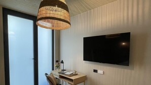 Een moderne kamer met een grote televisie aan de muur, een houten bureau, een stoel, een klein krukje en een groot raam met uitzicht.